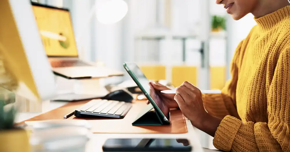 Person in a mustard yellow sweater uses a tablet with a stylus at a desk, with a keyboard and monitor nearby in a bright workspace.