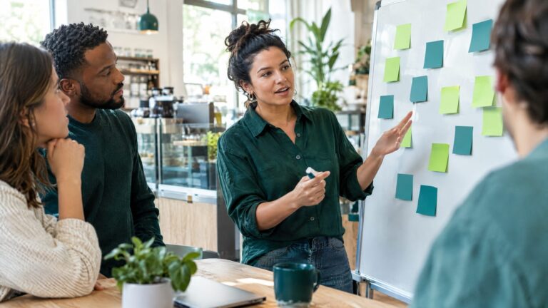 A small team planning growth at a whiteboard covered in sticky notes in lime and teal, a coffee shop owner in a deep green shirt leading